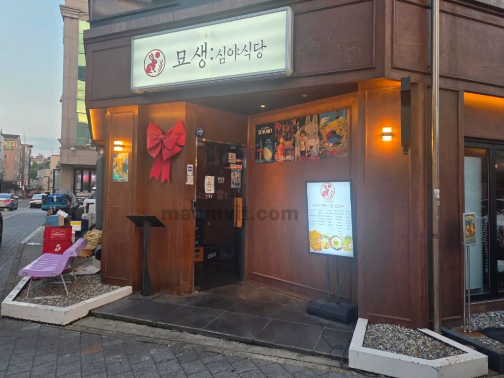 김포 장기동 맛집 묘생 심야식당은 먹자골목에 위치했어요. 따뜻한 우드 톤의 파사드와 은은한 조명, 커다란 빨간 리본 장식이 눈에 띄는 감성적인 선술집 입구입니다.